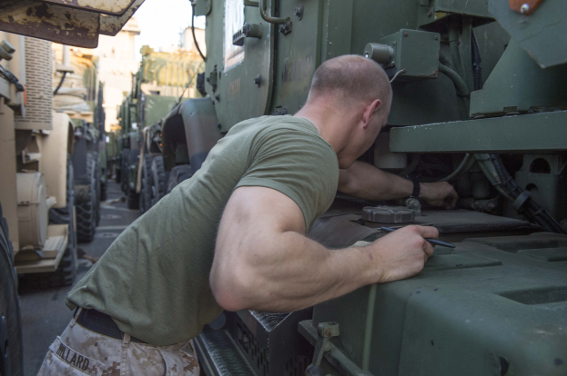 Ein Mann in militärischer Uniform repariert ein militärisches Fahrzeug auf einer Straße mit anderen Fahrzeugen, Gebäuden und einem klaren blauen Himmel im Hintergrund.