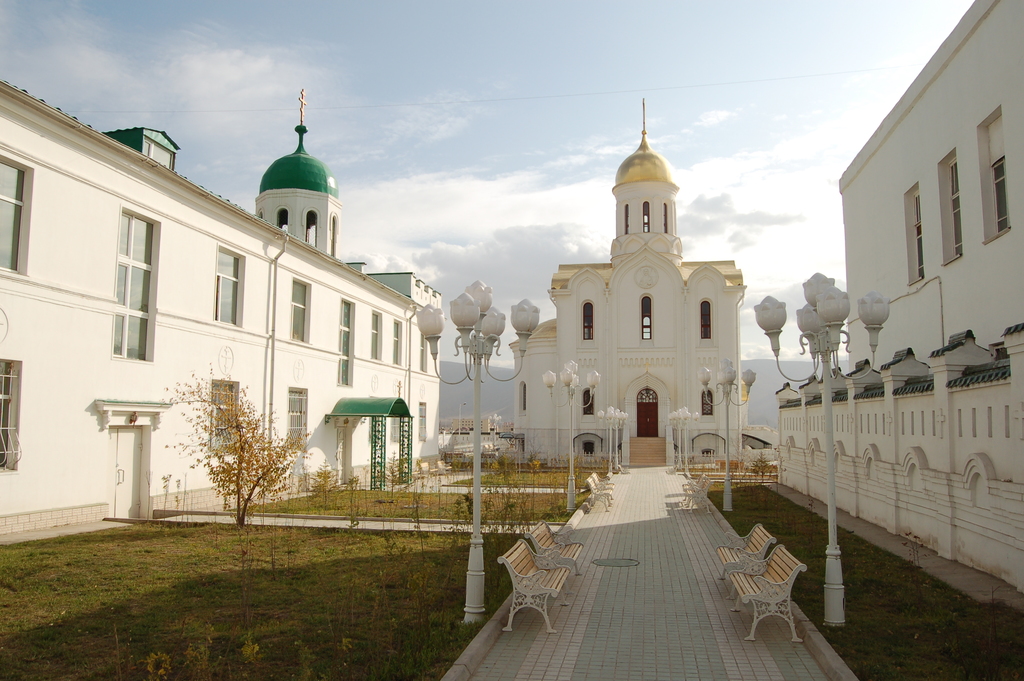 Eine Stadtansicht mit Gebäuden mit sichtbaren Fenstern, einer grünen Fläche mit einem Lichtmast, Blumenkübel, Bänken und einem Fußweg unter einem bewölkten Himmel.