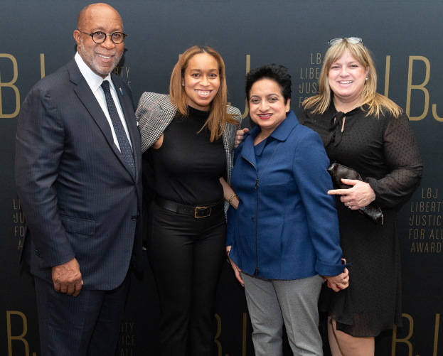 Eine Gruppe von vier Personen auf einem roten Teppich, die lächeln und für ein Foto posieren, mit einem 'Liberty Justice for All Awards' Schild im Hintergrund.