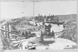Schwarzes und weißes Foto einer Baustelle mit einer Maschine im Vordergrund, umgeben von Hütten, Bäumen, Hügeln und Himmel im Hintergrund, und Text unten, der "Die Bauarbeiten an einer neuen Brücke in der Stadt San Francisco, Kalifornien" lautet.