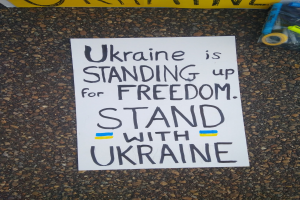 Weißer Zettel mit dem Text "Ukraine steht für Freiheit ein, steht mit Ukraine" auf dem Boden vor einer gelben Fahne, mit einer Plastikabdeckung, die verschiedene Gegenstände enthält, auf der rechten Seite.