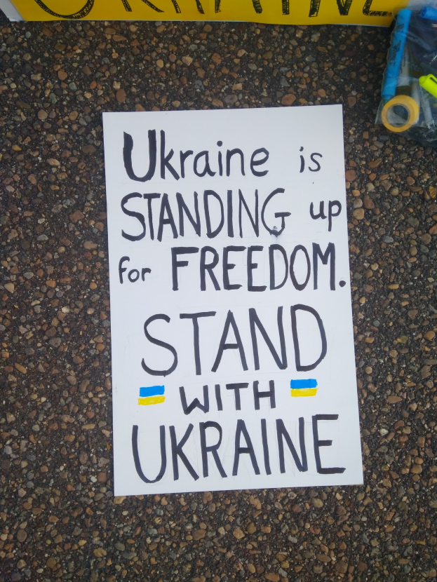 Weißer Zettel mit dem Text "Ukraine steht für Freiheit ein, steht mit Ukraine" auf dem Boden vor einer gelben Fahne, mit einer Plastikabdeckung, die verschiedene Gegenstände enthält, auf der rechten Seite.