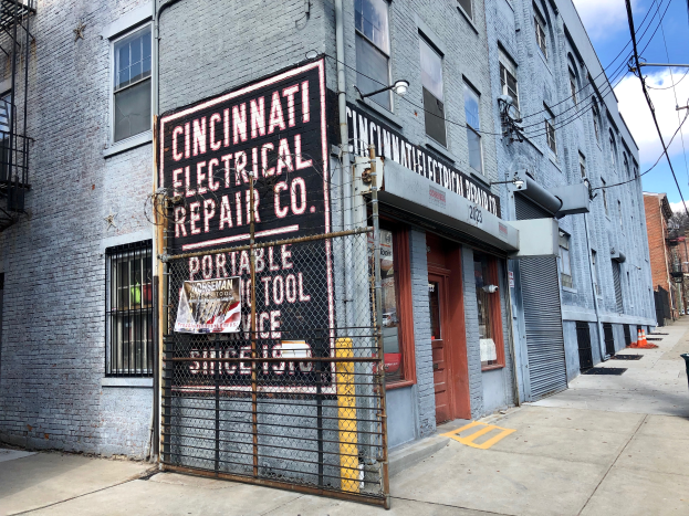 Das Cincinnati Electrical Repair Co. Gebäude mit Fenstern, Türen, einem Schild, einem Metallzaun, einem Müllcontainer, Pfählen, Drähten, Bäumen und einem bewölkten Himmel.