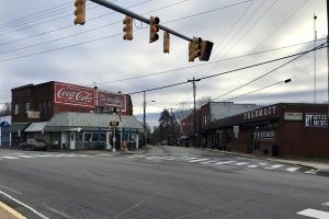 Stadtstraßenkreuzung mit Ampel, Fahrzeugen, Gebäuden mit Beschilderung, Strommasten mit Kabeln, Bäumen und einem bewölkten Himmel.