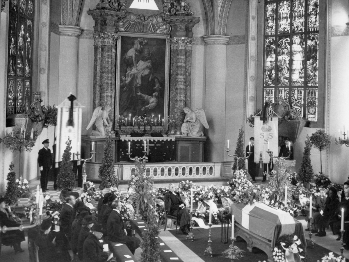 Ein Schwarz-Weiß-Foto einer Trauerfeier in einer Kirche, mit Sitzenden und Stehenden, Blumensträußen, Kerzen und einem Sarg im Vordergrund und einer Wand mit einem Foto, Skulpturen und Glasfenstern im Hintergrund.