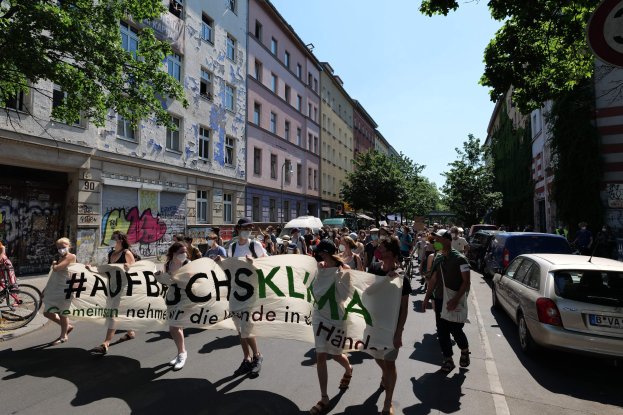 Eine Gruppe von Menschen marschiert auf einer Straße und trägt ein Banner, mit parkenden Autos auf der rechten Seite, einem Radfahrer auf der linken Seite, Gebäuden und Bäumen im Hintergrund und einem Schild, das auf eine Demonstration in Berlin hinweist.