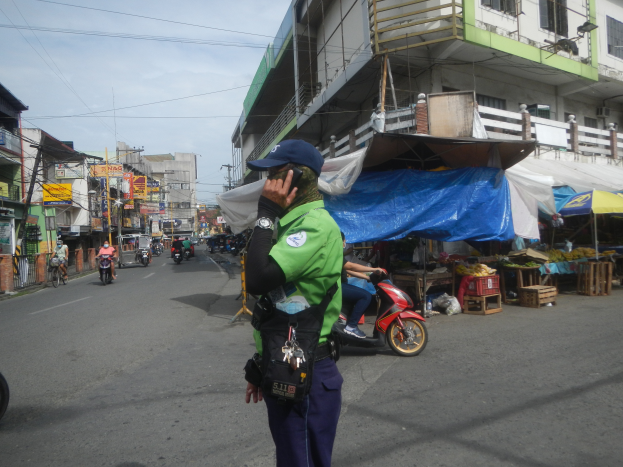 Ein Polizist in grüner und blauer Uniform und blauer Mütze steht am Straßenrand und spricht auf einem Handy, mit Fahrzeugen, die vorbeifahren, Ständen mit Gegenständen auf seiner rechten Seite und einem bewölkten Himmel im Hintergrund.