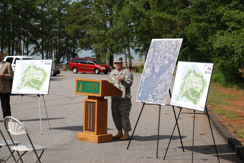 Eine Person steht an einem Podium mit einem Mikrofon, mit Plakaten, Stühlen, Fahrzeugen und Bäumen im Hintergrund.