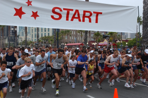 Gruppe von Läufern bei einem Marathon mit einem Verkehrskegel im Vordergrund und einem Banner im Hintergrund, umgeben von Bäumen, Laternenmasten, Gebäuden und einem klaren blauen Himmel.