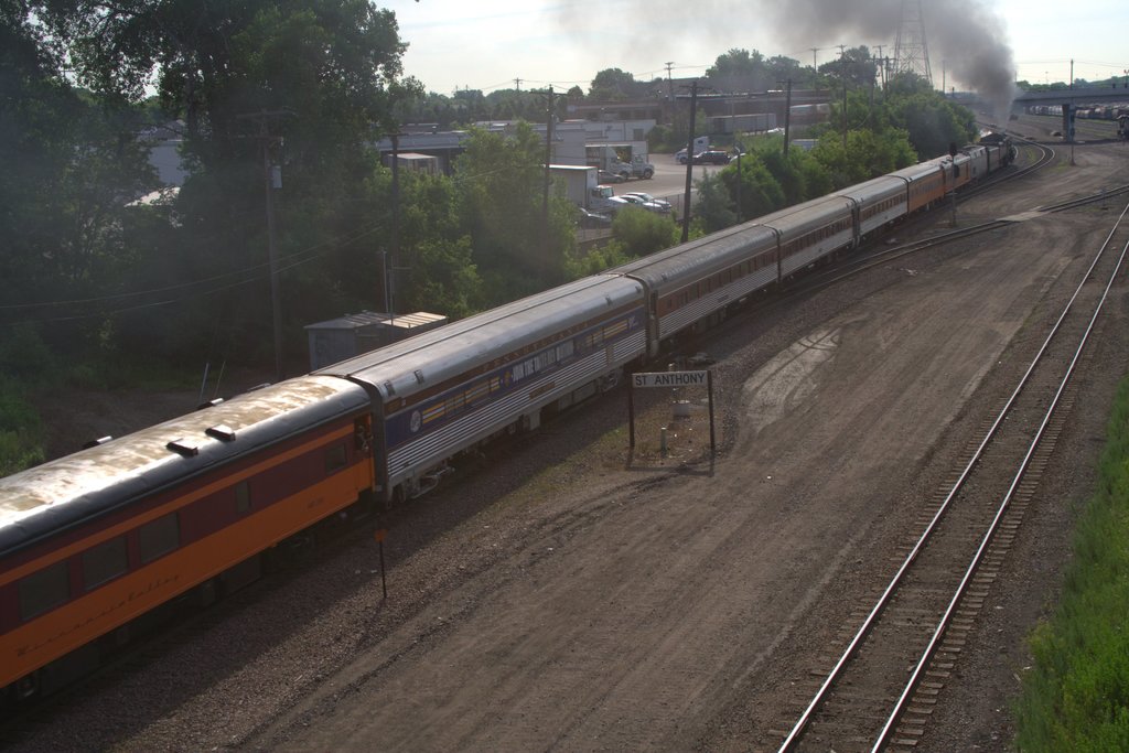 Ein Zug auf Schienen mit Bäumen, Strommasten mit Kabeln, Autos und Gebäuden links, eine Brücke davor und Rauch, der aus dem Zug aufsteigt.