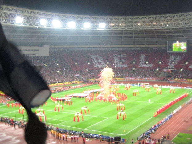 Ein Stadion mit Flutlicht, Eisengittern, Zuschauern, stehenden Personen, Werbetafeln, Anzeigetafeln, einer Sicherheitskamera, Fahnen und dekorativen Elementen.