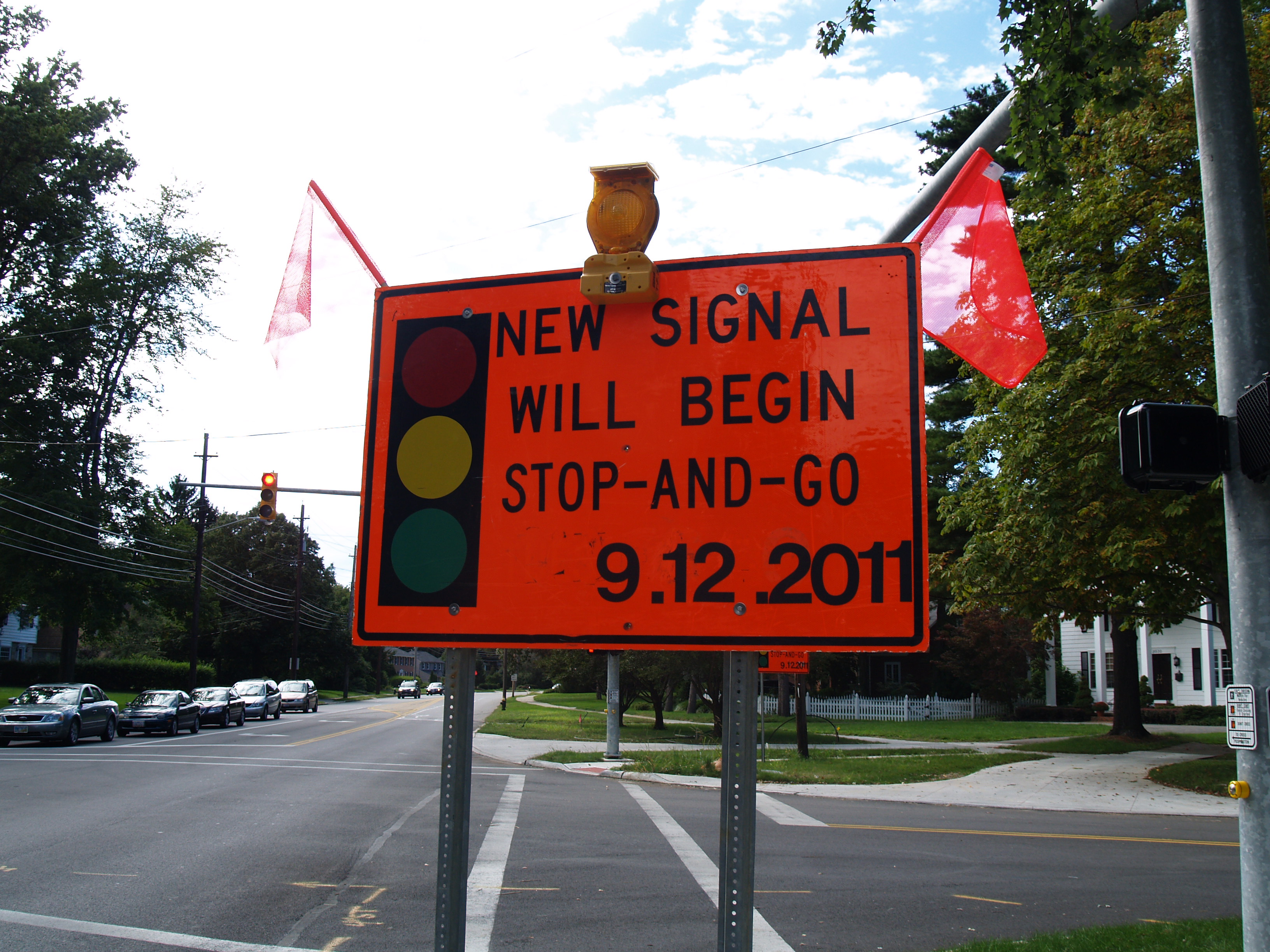 Eine Straße mit Fahrzeugen, ein Verkehrszeichen mit der Aufschrift "Neues Signal beginnt Stop and Go", Bäume, Gebäude und einen klaren blauen Himmel im Hintergrund.