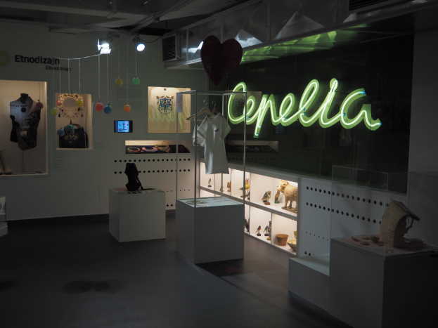 Ein Raum voller verschiedener Gegenstände, darunter Schaufensterpuppen mit Kleidern, Gegenstände auf Regalen, ein Display, ein Namensschild, dekorative Gegenstände an der Decke und Deckenleuchten bei der Cepella-Ausstellung in Mailand.