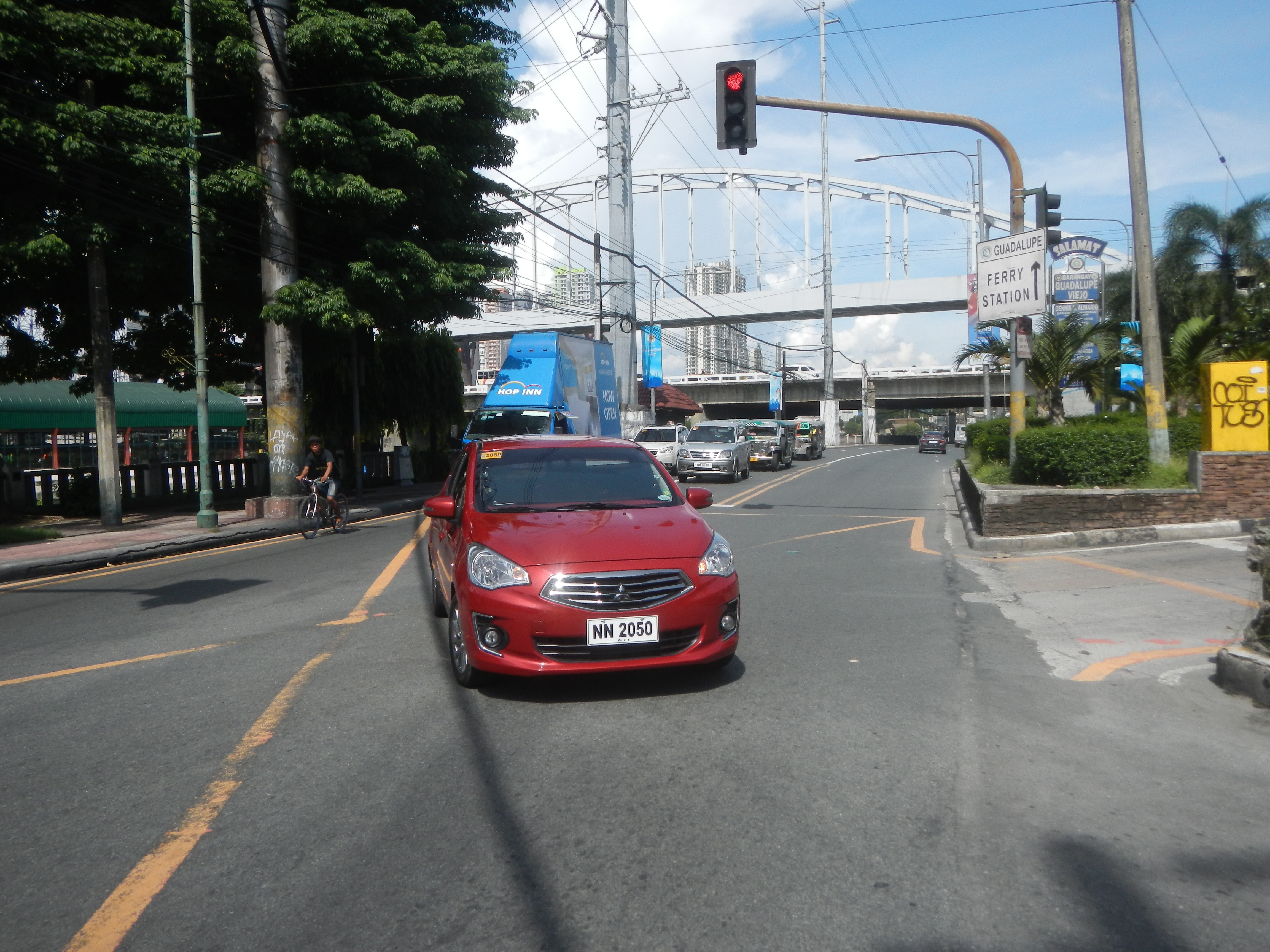 Ein rotes Auto fährt auf einer Straße neben einer Ampel, mit einem Radfahrer auf der linken Seite der Straße; im Hintergrund gibt es Bäume, Pfähle, Drähte, eine Brücke, Gebäude und einen bewölkten Himmel.