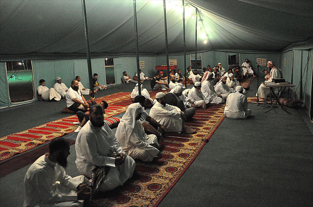 Eine Gruppe von Menschen sitzt auf dem Boden in einem Lager, mit einer Person auf einem Stuhl rechts, einem Mikrofon vor ihnen, einem Tisch neben ihnen und einer Lampe oben im Bild.