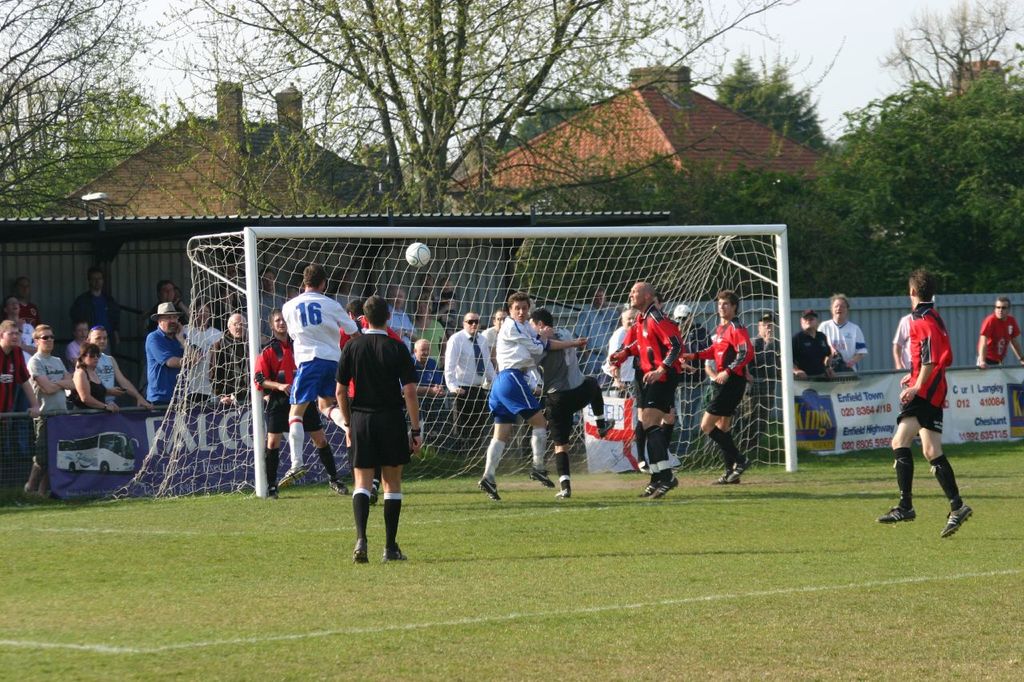 Spieler spielen Fußball auf einem Feld mit einem Tor dahinter, während Zuschauer von der Seitenlinie zuschauen, mit Bäumen und Häusern im Hintergrund.