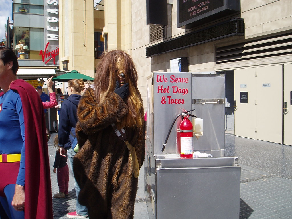 Eine Menge Menschen, einige in Kostümen, versammelten sich vor Gebäuden mit sichtbarer Beschriftung, einer Box mit Schrift und einem Feuerlöscher.