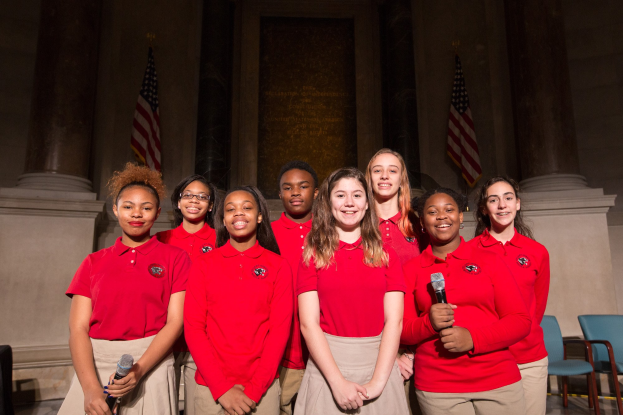 Gruppe junger Frauen in roten Shirts, die strahlend Mikrofone halten und vor Stühlen, Säulen, Flaggen und einer Wand stehen, was an eine nationale Jugendchor-Aufführung denken lässt.