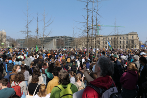 Eine große Menge von Menschen mit Schildern und Taschen steht vor einem Gebäude mit Bäumen und einem klaren Himmel während einer Klimaprotest.