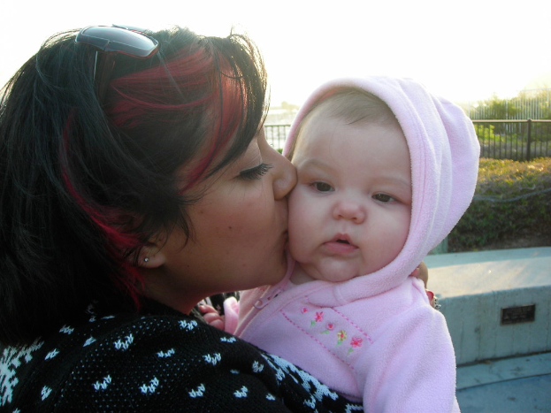 Eine Frau hält ein Baby in der Mitte des Bildes, mit Büschen und Zaun im Hintergrund und Boden sichtbar in der rechten unteren Ecke.