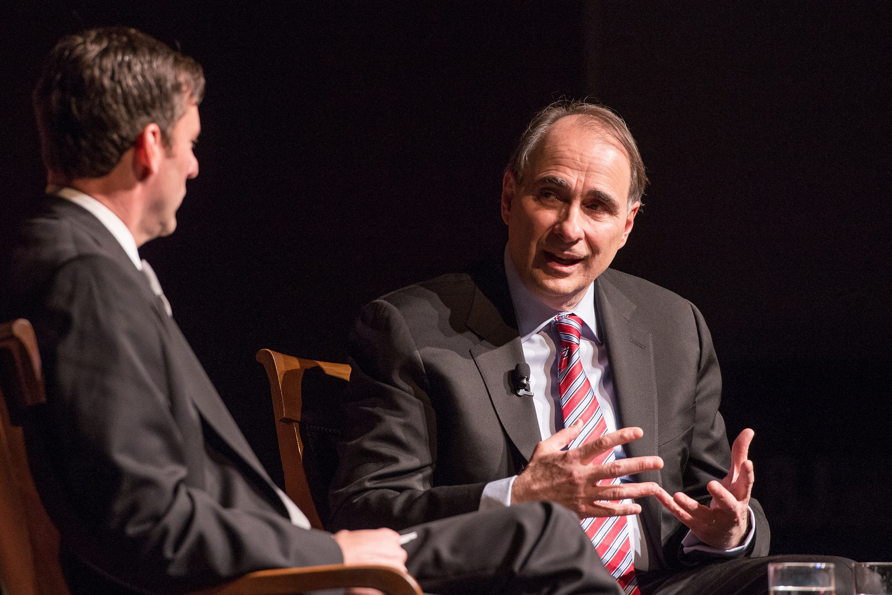 Zwei Männer in Anzügen sitzen sich in Stühlen gegenüber und unterhalten sich mit einem Glas Wasser auf dem Tisch dazwischen.