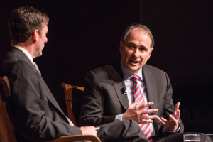 Zwei Männer in Anzügen sitzen sich in Stühlen gegenüber und unterhalten sich mit einem Glas Wasser auf dem Tisch dazwischen.