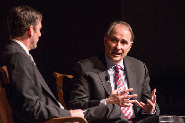 Zwei Männer in Anzügen sitzen sich in Stühlen gegenüber und unterhalten sich mit einem Glas Wasser auf dem Tisch dazwischen.