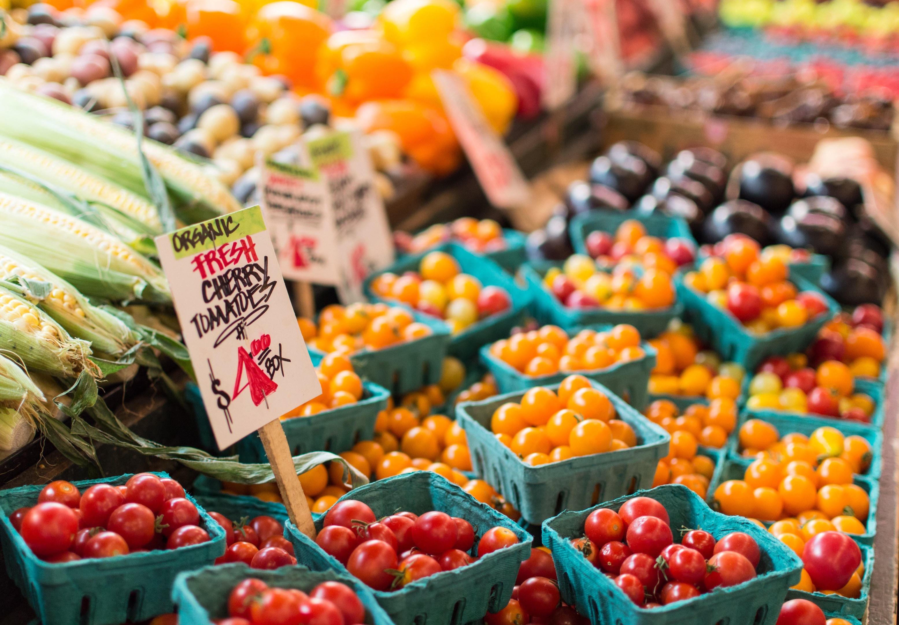 Frisches Obst und Gemüse, darunter Tomaten, Mais und andere Gemüse, in Körben auf einem Bauernmarkt mit Texttafeln im Hintergrund.