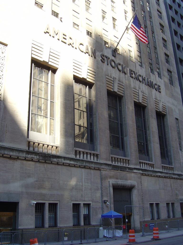 Ein Gebäude mit Text an seiner Fassade, eine Fahnenstange an der Wand, ein Geländer auf dem Gehweg davor, Absperrungen jenseits des Geländers und eine Straße im rechten unteren Bereich.