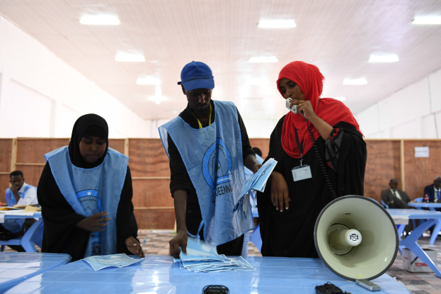 Eine Gruppe von Menschen an einem Wahlurnentisch mit einem Megafon, Papieren und anderen Gegenständen, während andere im Hintergrund unter beleuchteter Decke sitzen und stehen.