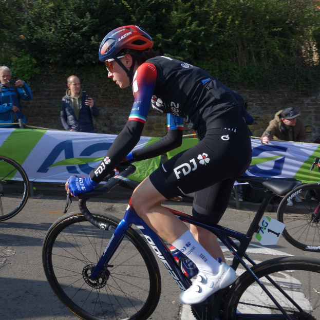 Eine professionelle Radrennfahrerin, Katie Archibald, trägt einen Helm, eine Brille, Handschuhe und Schuhe und fährt ein Fahrrad auf einer Straße neben einer Menschenmenge, mit einem Banner, einer Wand, Bäumen und einem Gebäude im Hintergrund.