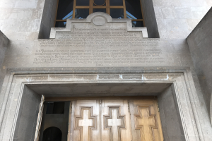 Große hölzerne Tür vor einem Steinbau mit einem Glasfenster oben, mit Inschrift auf der Wand, die wahrscheinlich Holocaust-Opfer gedenkt.