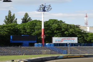 Rennstrecke mit Flutlicht, umgeben von Gras, Geländern, Stufen, einem Lichtmast, einem Schild, Bäumen, Gebäuden, Türmen und einem bewölkten Himmel.