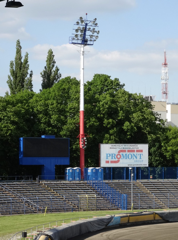 Rennstrecke mit Flutlicht, umgeben von Gras, Geländern, Stufen, einem Lichtmast, einem Schild, Bäumen, Gebäuden, Türmen und einem bewölkten Himmel.