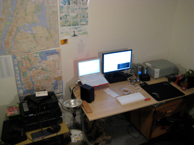 Ein Raum mit einem Tisch, auf dem ein Laptop, ein Lautsprecher, ein Monitor, eine Tastatur, ein Ball, eine Maus, ein Scanner und Papiere stehen, sowie ein weiterer Tisch mit elektronischen Geräten und Charts und Poster an der Wand dahinter.