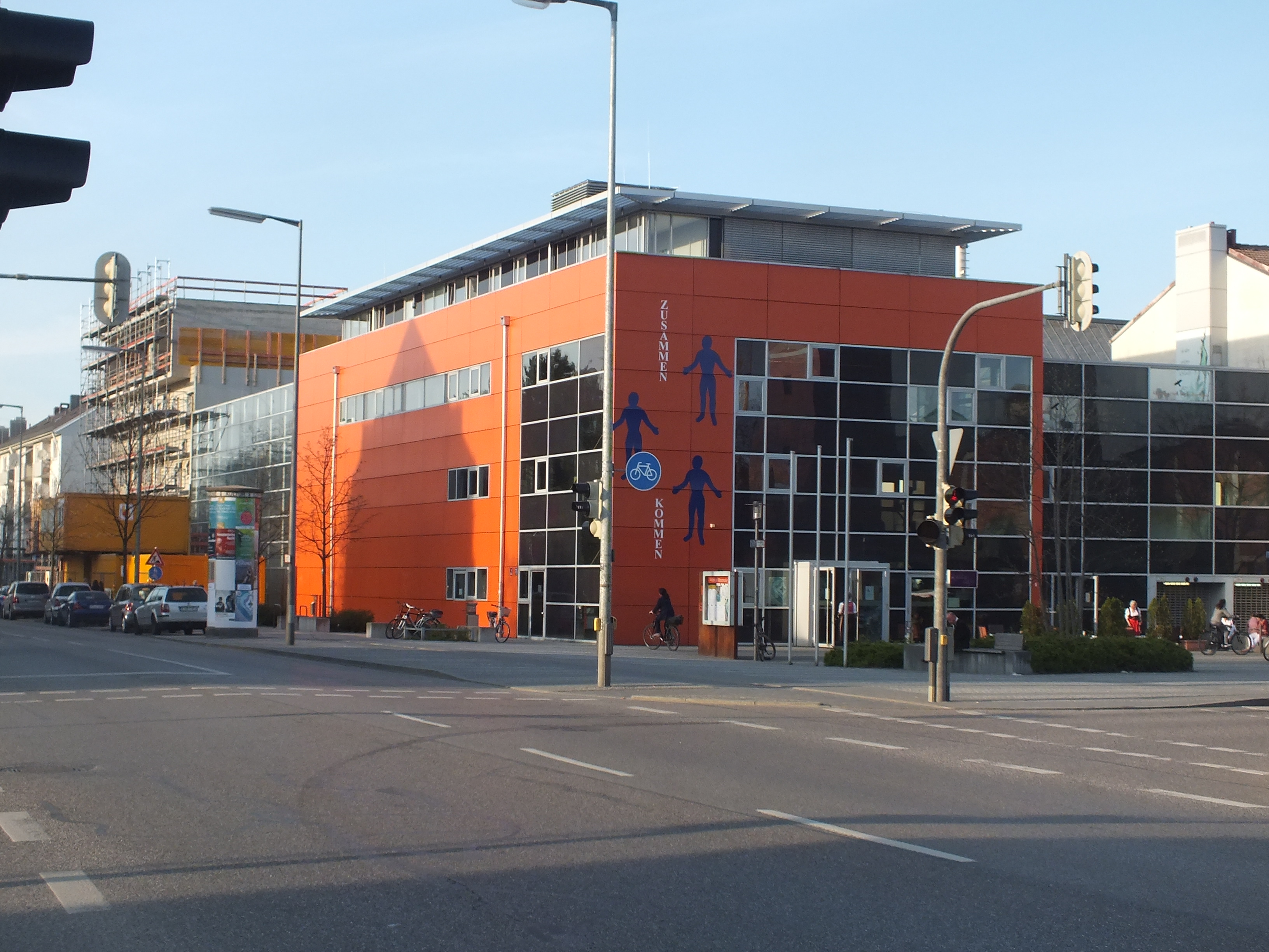 Ein gro├če orange Geb├Ąude an einer Stra├čenkreuzung mit Fahrzeugen, Radfahrern, Stra├čenmasten, Lichtern und Signalen, umgeben von B├Ąumen und B├╝schen unter einem bew├Âlktem Himmel.