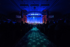 Ein großer Saal mit sitzenden Zuschauern, eine Bühne mit Musikern und Deckenbeleuchtung und Bildschirme, was auf ein Konzerthaus während der Nationalkonvention 2019 hinweist.