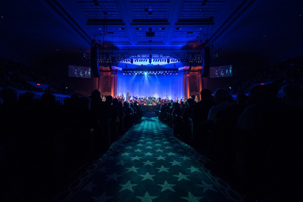 Ein großer Saal mit sitzenden Zuschauern, eine Bühne mit Musikern und Deckenbeleuchtung und Bildschirme, was auf ein Konzerthaus während der Nationalkonvention 2019 hinweist.