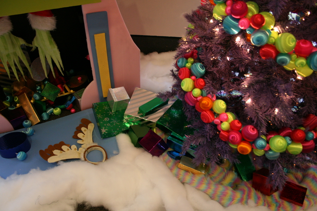 Drinnen steht ein geschm├╝ckter Weihnachtsbaum mit Lichterkette und Schmuck rechts, mit Geschenkboxen und Deko in der Mitte, eine H├╝tte mit Deko oben links und Baumwolle und Wolle unten.