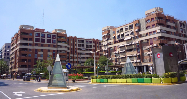 Eine Stadtstraße mit einem zentralen Denkmal, hohen Gebäuden, Straßenmöblierung, Grünflächen, Fußgängern und einem klaren blauen Himmel.