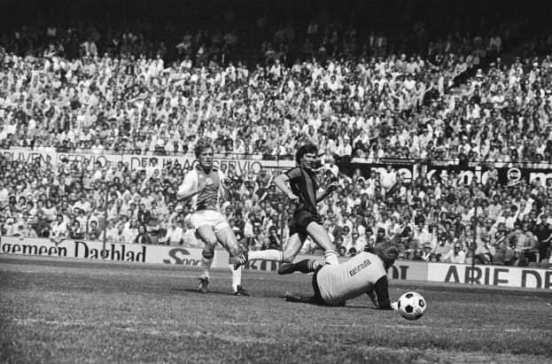 Schwarz-weiß-Foto von Männern, die auf einem Feld Fussball spielen, mit Zuschauern im Hintergrund und einer "Bundesliga 1978-79 Ajax 1-1 Hannover 96"-Tafel.