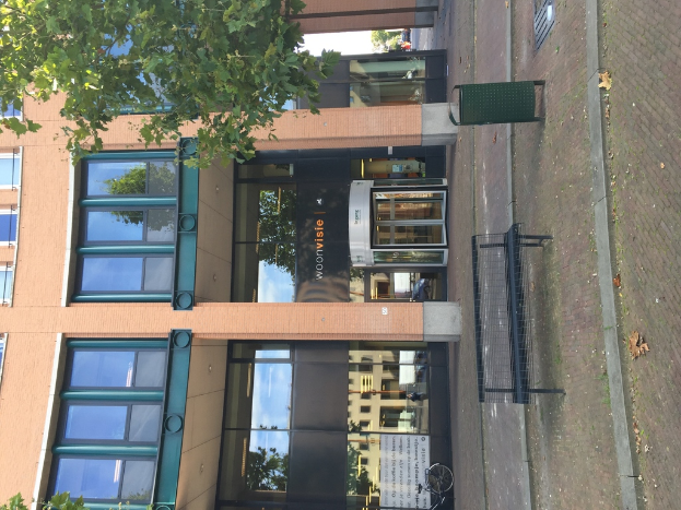 Ein Geb√§ude mit einem M√ľllimer, Bank, Baum und Banner davor.