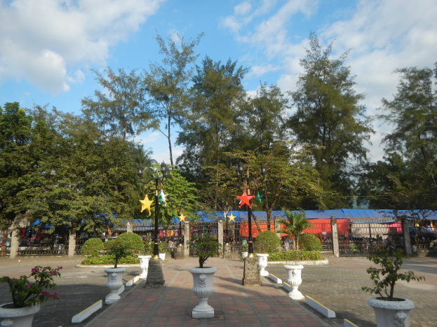 Ein Park mit Topfpflanzen und Bäumen in der Mitte, umgeben von Straßenlaternen, Dekorationsgegenständen und Grills unter einem bewölkten Himmel.