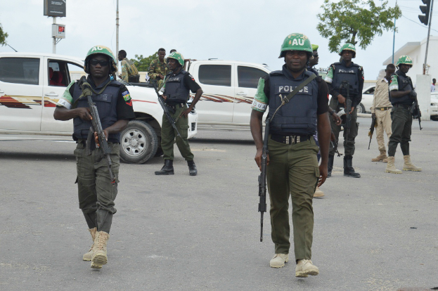Eine Gruppe von Polizisten in Helmen und mit Waffen in Händen geht eine Straße entlang mit Fahrzeugen, Laternenmasten, Ampeln, Texttafeln, Bäumen, Gebäuden und einem bewölktem Himmel im Hintergrund.