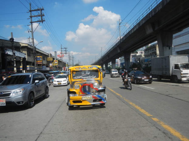 Gelbes Taxi fährt auf einer Straße mit hohen Gebäuden, Strommasten mit Drähten und einer Brücke im Hintergrund bei bewölktem Himmel mit einem Wasserzeichen.