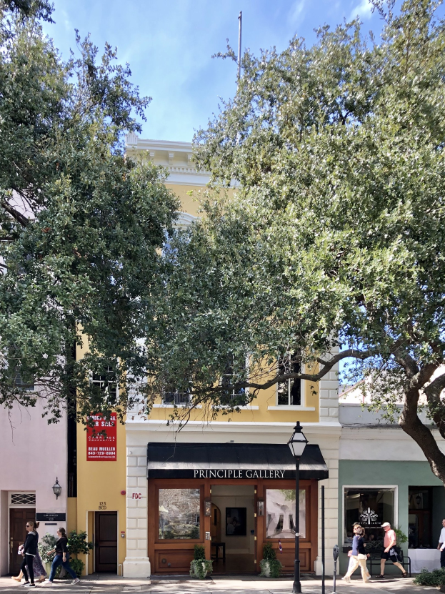 Außenansicht der Principle Gallery in Charleston, SC, mit Menschen auf dem Gehweg, Topfpflanzen, einer Straßenlaterne, einem Schild, einer Fahne mit Text und einem Wolkenhimmel.