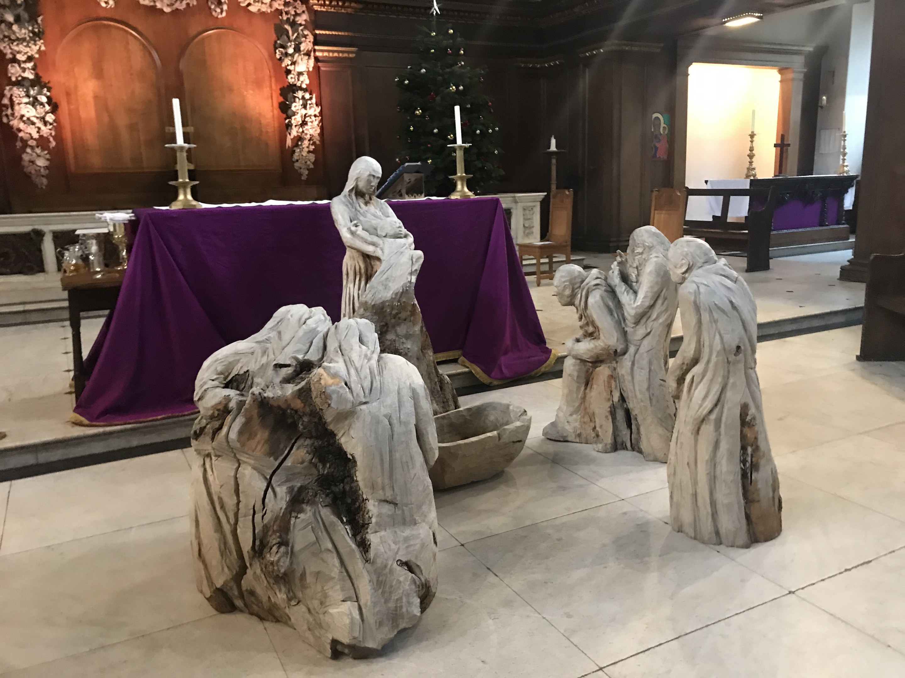 Krippe in einer Kirche mit Statuen, einem geschmückten Weihnachtsbaum, Stühlen, einem Tisch mit Kerzen und einem tischgedeckten Tisch.