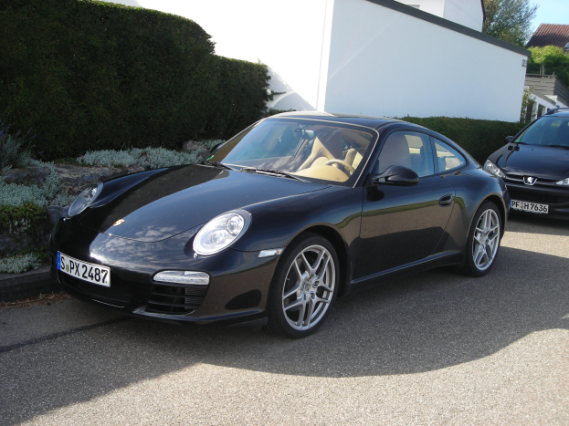 Zwei schwarze Porsche 911 vor einem weißen Haus geparkt, umgeben von grünen Pflanzen und Bäumen, mit einem hellblauen Himmel im Hintergrund.