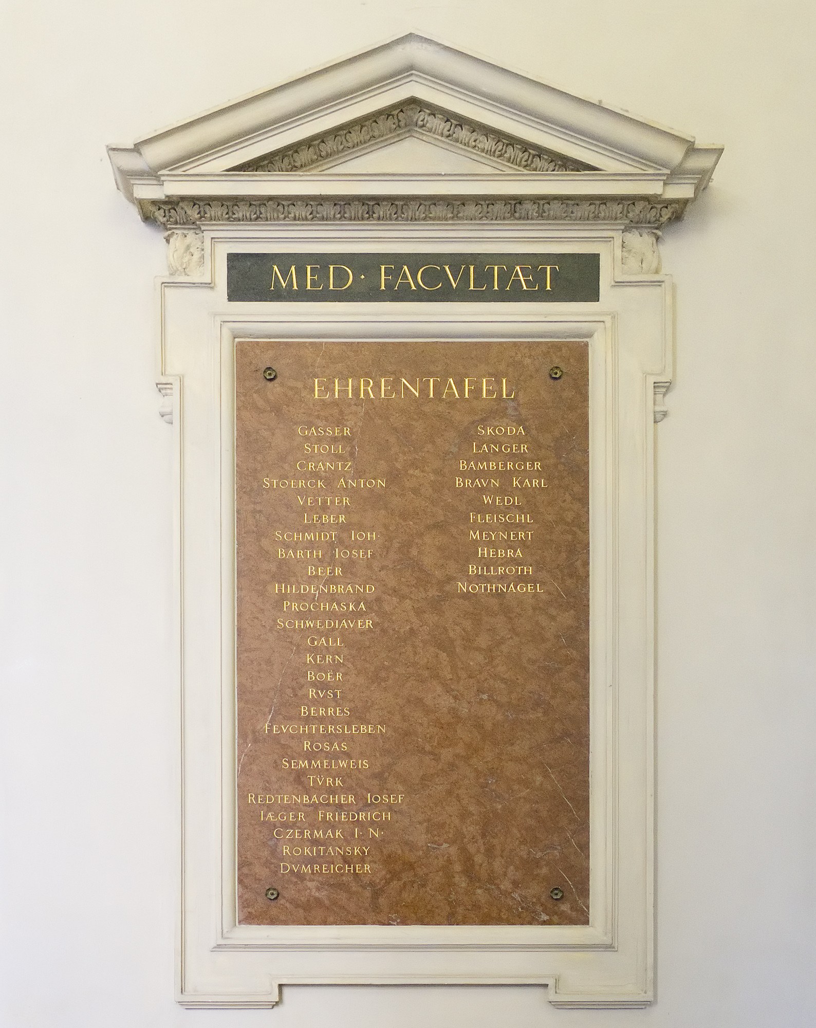 Weißer rechteckiger Schild an einer Gebäudewand mit schwarzer fettgedruckter Schrift, die "Med Facilitat" in der Mitte liest.