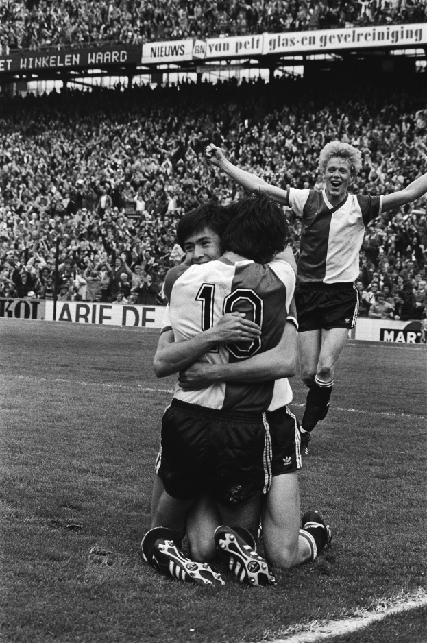 Drei Fussballspieler umarmen sich auf dem Boden und feiern ein Tor vor einer jubelnden Menge in einer schwarz-weißen Stadionszene.